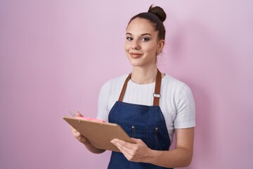 Young hispanic girl wearing professional waitress apron taking order smiling with a happy and cool...