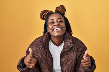 African woman with braided hair standing over yellow background success sign doing positive gesture...