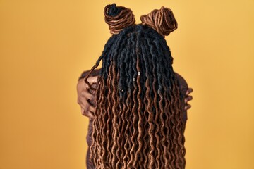 African woman with braided hair standing over yellow background hugging oneself happy and positive from backwards. self love and self care - obrazy, fototapety, plakaty