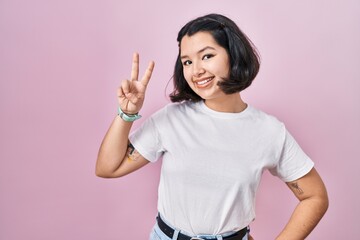 Young hispanic woman wearing casual white t shirt over pink background smiling looking to the camera showing fingers doing victory sign. number two.