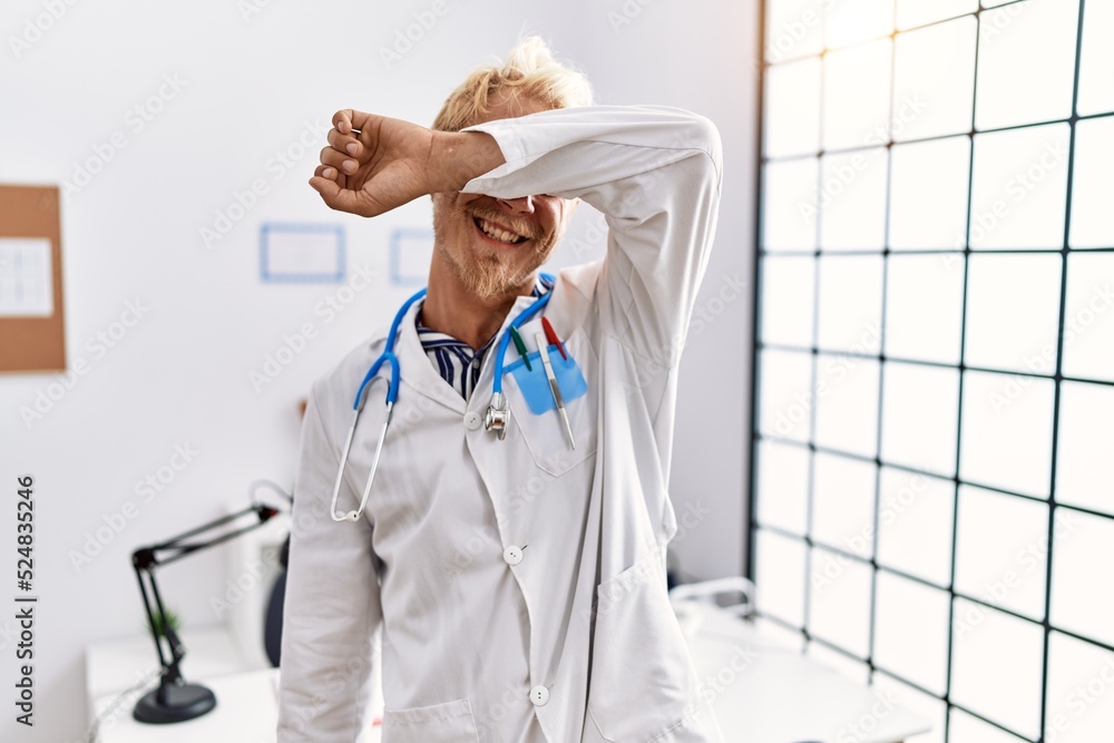 Sticker Young blond man wearing doctor uniform and stethoscope at clinic smiling cheerful playing peek a boo with hands showing face. surprised and exited