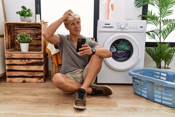 Young blond man doing laundry using smartphone confuse and wondering about question. uncertain with...