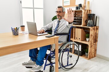 Middle age hispanic man working at the office sitting on wheelchair doing stop sing with palm of the hand. warning expression with negative and serious gesture on the face.