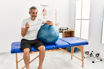 Middle age hispanic man at pain recovery clinic holding pilates ball doing money gesture with...