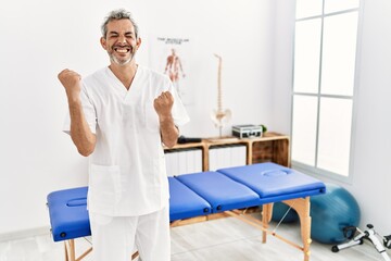 Fototapeta premium Middle age hispanic therapist man working at pain recovery clinic very happy and excited doing winner gesture with arms raised, smiling and screaming for success. celebration concept.