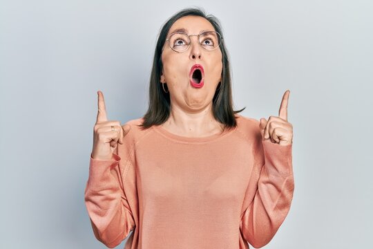 Middle age hispanic woman wearing casual clothes and glasses amazed and surprised looking up and pointing with fingers and raised arms.