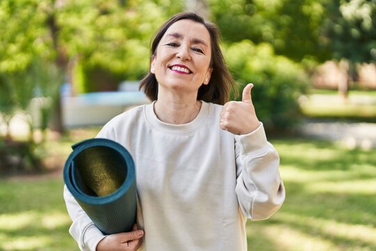 Middle age hispanic woman holding yoga mat at the park smiling happy and positive, thumb up doing excellent and approval sign