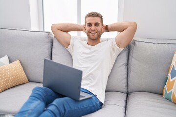 Young caucasian man using laptop relaxed with hands on head at home