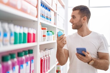 Young caucasian man customer using smartphone holding deodorant at pharmacy