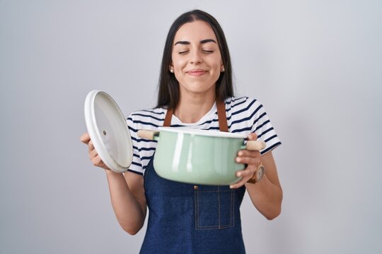 Young brunette woman wearing apron holding cooking pot smiling looking to the side and staring away thinking.
