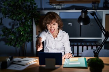 Middle age woman working at the office at night pointing displeased and frustrated to the camera,...