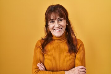 Middle age hispanic woman standing over yellow background happy face smiling with crossed arms looking at the camera. positive person.