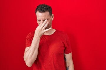 Young hispanic man standing over red background smelling something stinky and disgusting, intolerable smell, holding breath with fingers on nose. bad smell