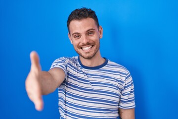 Fototapeta premium Young hispanic man standing over blue background smiling friendly offering handshake as greeting and welcoming. successful business.