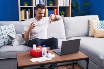Young hispanic man watching video on laptop on how to use first aid kit with angry face, negative sign showing dislike with thumbs down, rejection concept