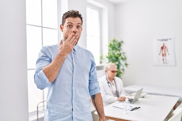 Hispanic man at doctor clinic covering mouth with hand, shocked and afraid for mistake. surprised...
