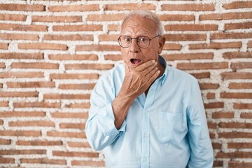 Senior man with grey hair standing over bricks wall looking fascinated with disbelief, surprise and...