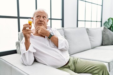 Senior man holding pills thinking worried about a question, concerned and nervous with hand on chin