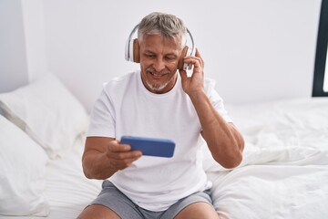 Middle age grey-haired man listening to music sitting on bed at bedroom