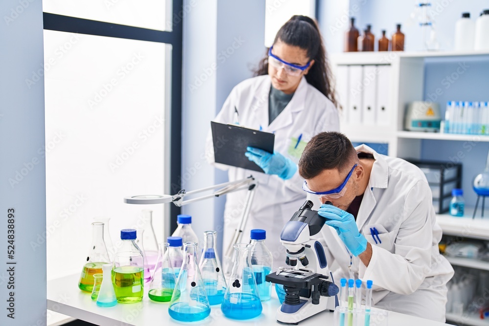 Wall mural Man and woman wearing scientist uniform using microscope write on document at laboratory