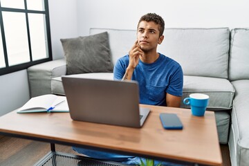Young handsome hispanic man using laptop sitting on the floor with hand on chin thinking about question, pensive expression. smiling with thoughtful face. doubt concept.