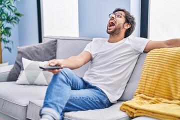 Handsome latin man holding television remote control angry and mad screaming frustrated and furious, shouting with anger looking up.