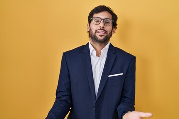 Handsome latin man standing over yellow background smiling cheerful with open arms as friendly welcome, positive and confident greetings