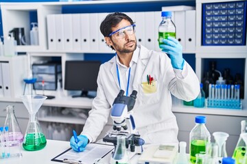 Young hispanic man scientist write on clipboard holding bottle at laboratory