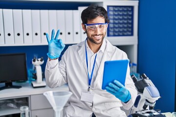 Handsome latin man working at scientist laboratory using tablet doing ok sign with fingers, smiling friendly gesturing excellent symbol