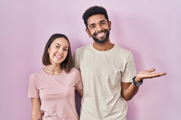 Young hispanic couple together over pink background smiling cheerful presenting and pointing with palm of hand looking at the camera.