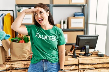 Young brunette woman wearing volunteer t shirt at donations stand very happy and smiling looking far away with hand over head. searching concept.