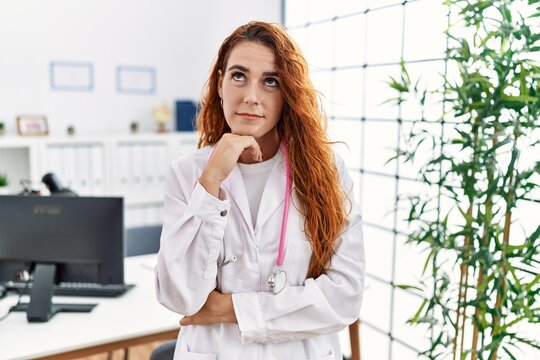 Young redhead woman wearing doctor uniform and stethoscope at the clinic with hand on chin thinking about question, pensive expression. smiling with thoughtful face. doubt concept.