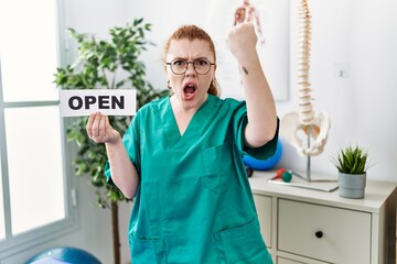 Young redhead physiotherapist woman working at pain recovery clinic holding open banner annoyed and frustrated shouting with anger, yelling crazy with anger and hand raised