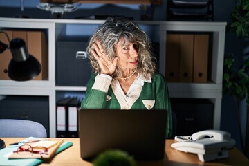 Middle age woman working at night using computer laptop smiling with hand over ear listening an hearing to rumor or gossip. deafness concept.