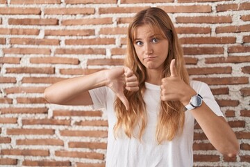 Young caucasian woman standing over bricks wall doing thumbs up and down, disagreement and agreement expression. crazy conflict