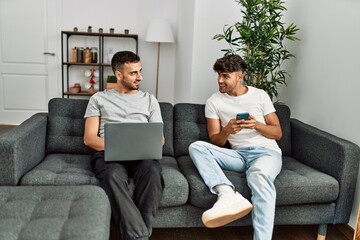 Two hispanic men couple using smartphone and laptop at home