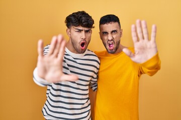 Young hispanic gay couple standing over yellow background doing stop gesture with hands palms, angry and frustration expression
