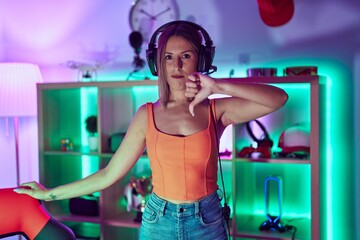 Young woman playing video games wearing headphones with angry face, negative sign showing dislike with thumbs down, rejection concept