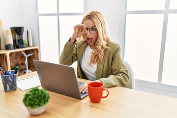 Beautiful blonde woman working at the office with laptop doing ok gesture shocked with surprised face, eye looking through fingers. unbelieving expression.