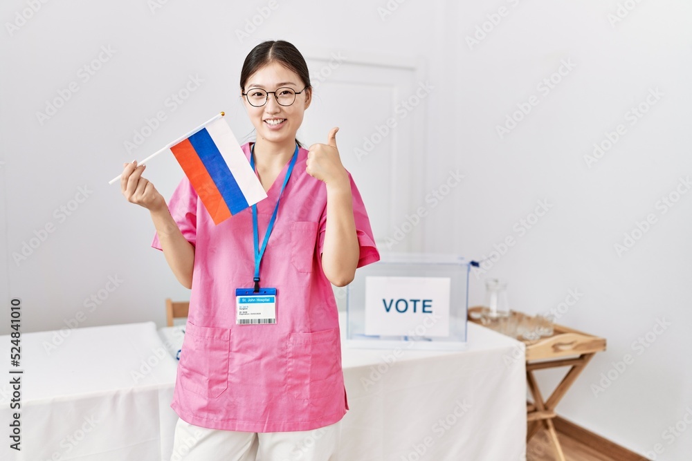 Sticker Young asian nurse woman at political campaign election holding russia flag smiling happy and positive, thumb up doing excellent and approval sign