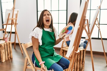 Young hispanic artist women painting on canvas at art studio angry and mad screaming frustrated and furious, shouting with anger. rage and aggressive concept.