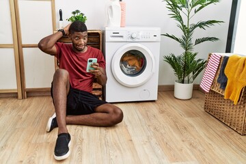 Young african american man using smartphone waiting for washing machine crazy and scared with hands...