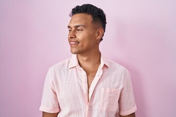 Young hispanic man standing over pink background looking away to side with smile on face, natural expression. laughing confident.