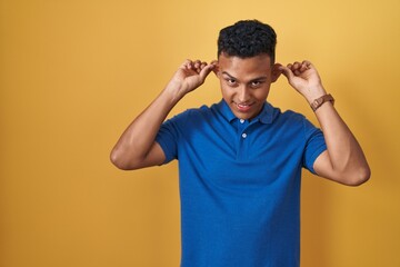 Young hispanic man standing over yellow background smiling pulling ears with fingers, funny...