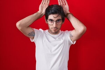 Young hispanic man standing over red background doing bunny ears gesture with hands palms looking cynical and skeptical. easter rabbit concept.