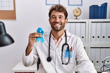 Young hispanic doctor man holding blue ribbon looking positive and happy standing and smiling with...