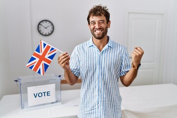 Young handsome man at political campaign election holding uk flag screaming proud, celebrating victory and success very excited with raised arm