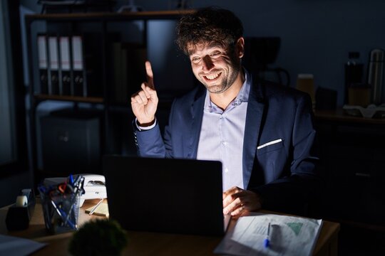 Hispanic young man working at the office at night pointing finger up with successful idea. exited and happy. number one.