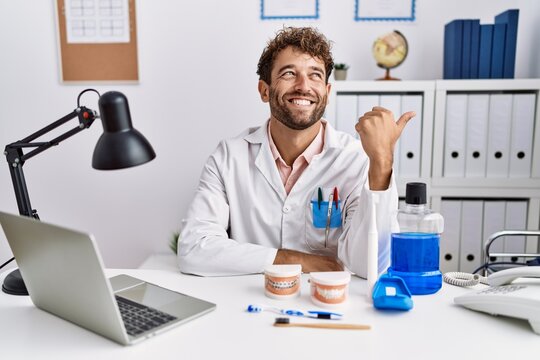 Young hispanic dentist man working at medical clinic smiling with happy face looking and pointing to the side with thumb up.