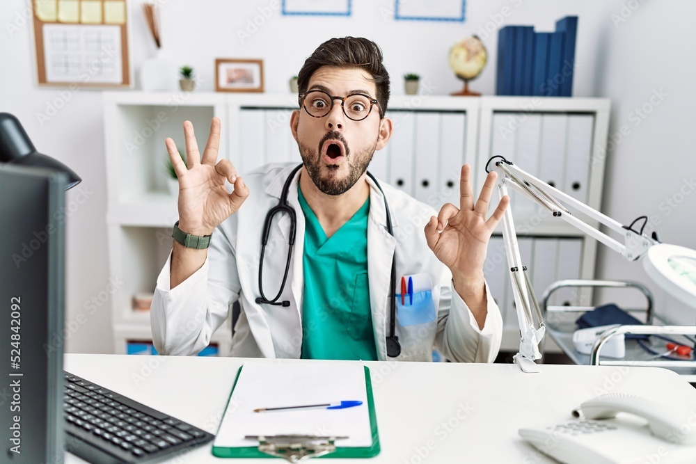 Poster young man with beard wearing doctor uniform and stethoscope at the clinic looking surprised and shoc